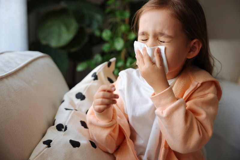 A young girl blowing or wiping her nose with a tissue while sitting on a couch indoors.